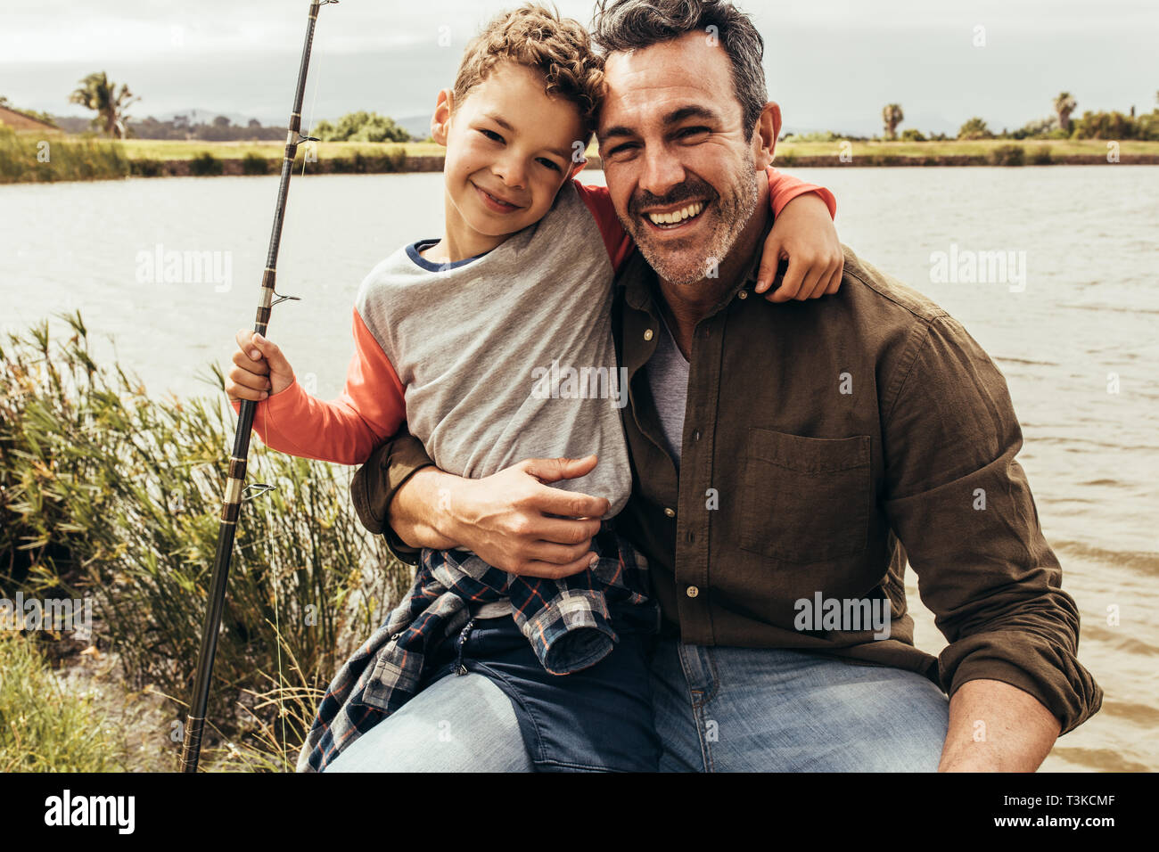 Smiling father and son sitting beside a lake holding a fishing rod. Close up of father and son on a day out spending time together fishing near a lake Stock Photo