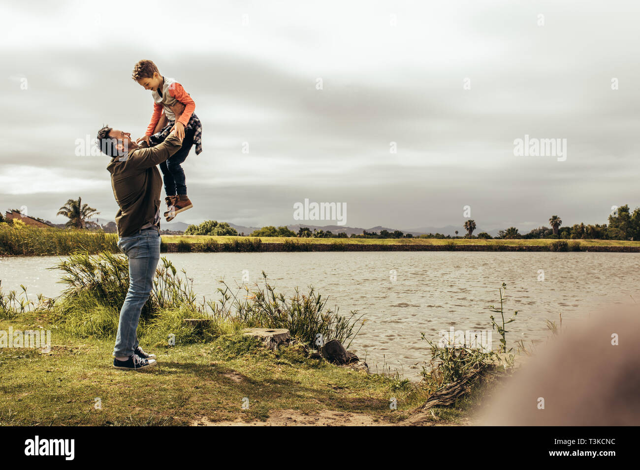 Father and son having fun spending time together outdoors. Man lifting his son high playing with him standing near a lake. Stock Photo