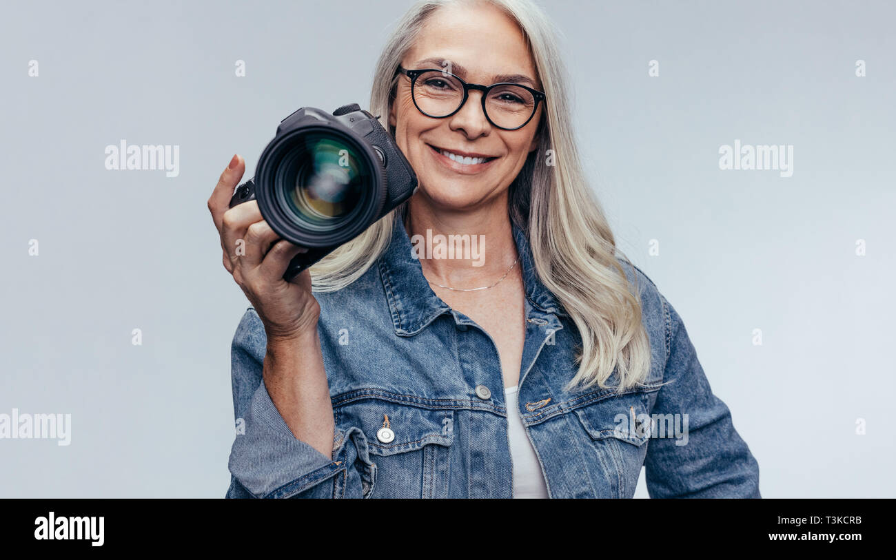 Confident senior woman in casuals holding a dslr camera on grey background. Professional female photographer with digital camera. Stock Photo