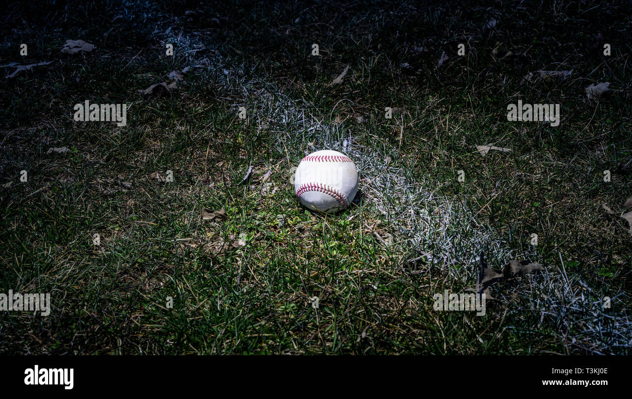 Single Baseball in outfield grass visibly next to chalk line in low light with light shining on