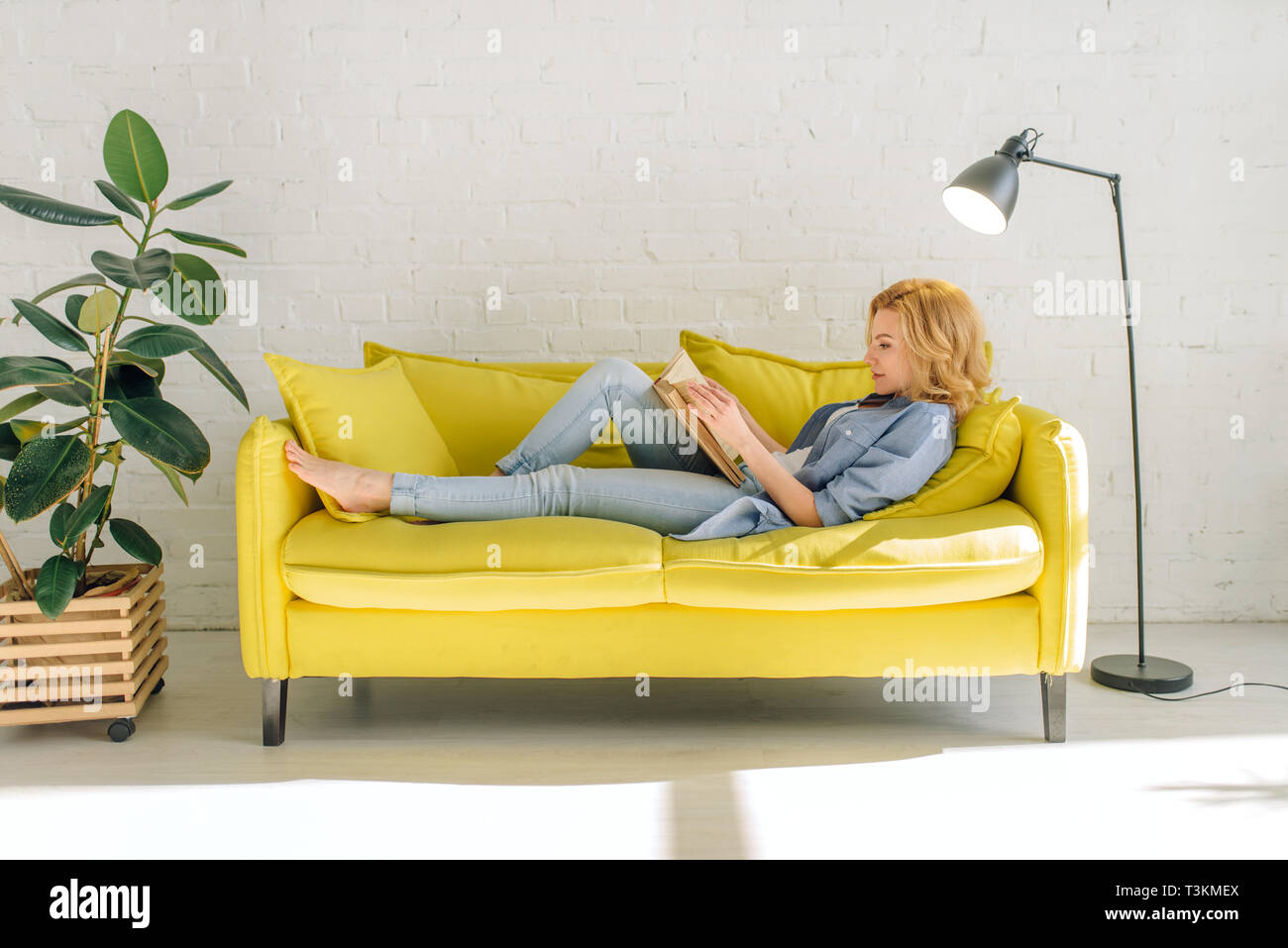 Woman Lying On Cozy Yellow Couch And Reading Book Stock Photo
