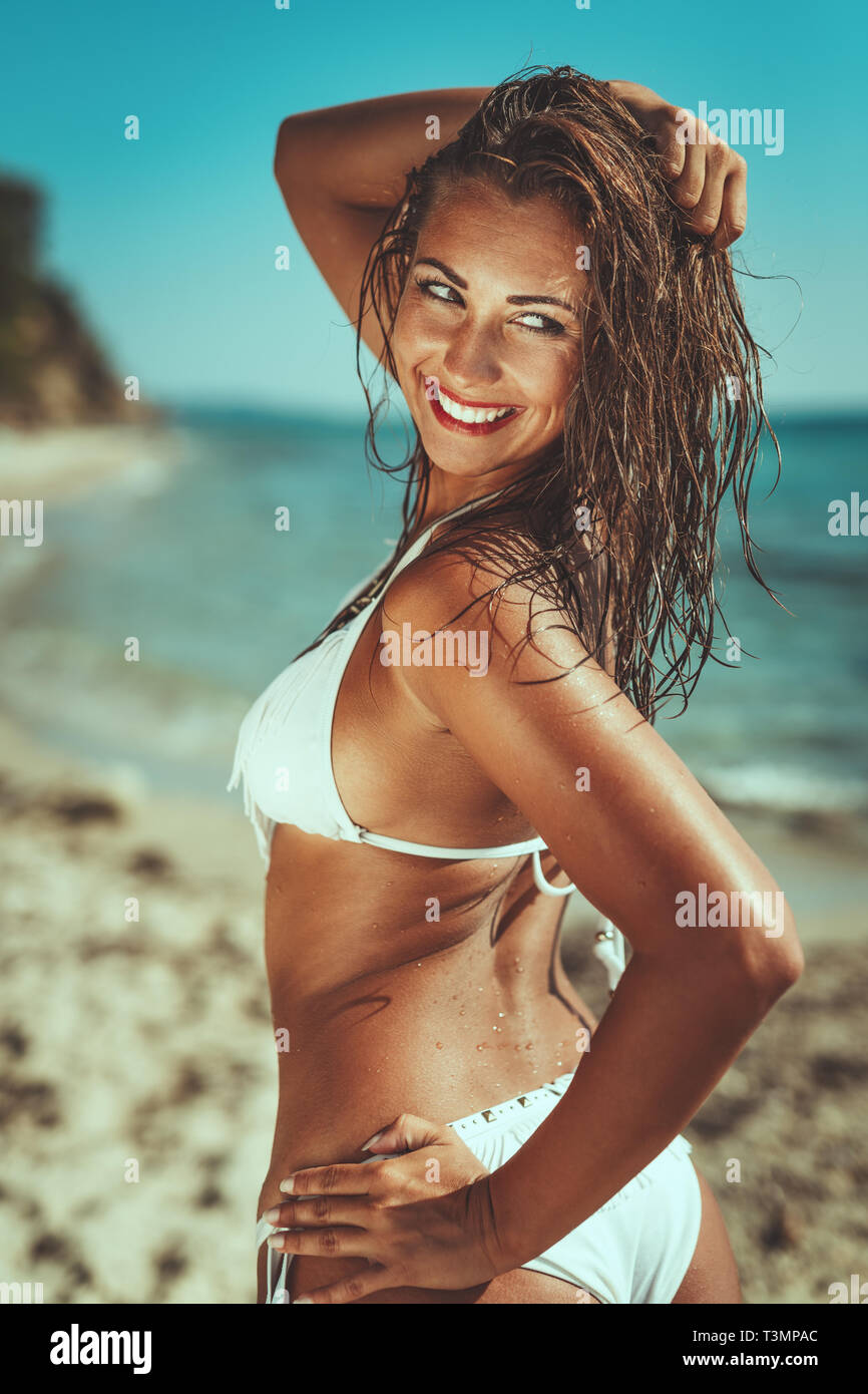 A beautiful young woman in swimsuit is enjoying at sunset on the beach with raised arm. Stock Photo