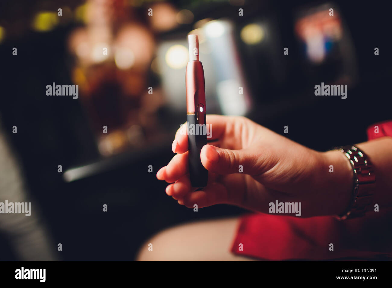 Electronic cigarettes, technology cigarette. Tobacco system IQOS Female hand on blurred background of the restaurant. red case. Stock Photo