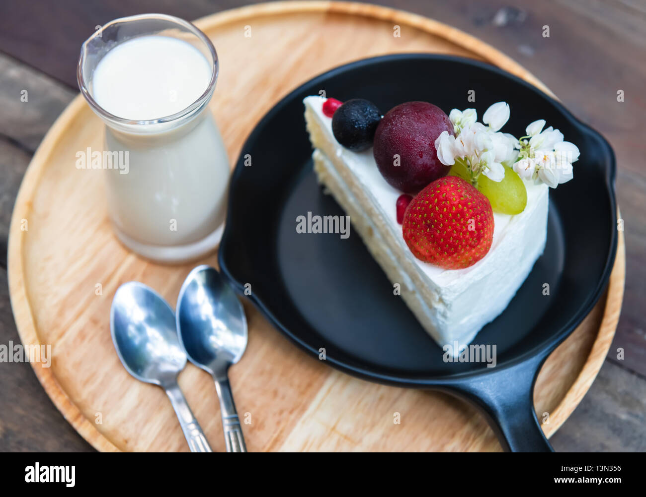 Sweet fruit cream cake cut serve with milk glass set on wooden plate with indoor low lighting. Stock Photo