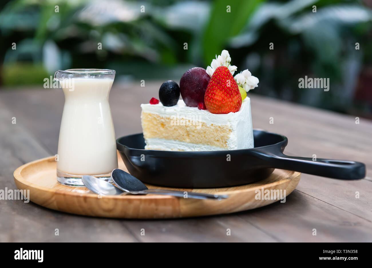 Sweet fruit cream cake cut serve with milk glass set on wooden plate with indoor low lighting. Stock Photo