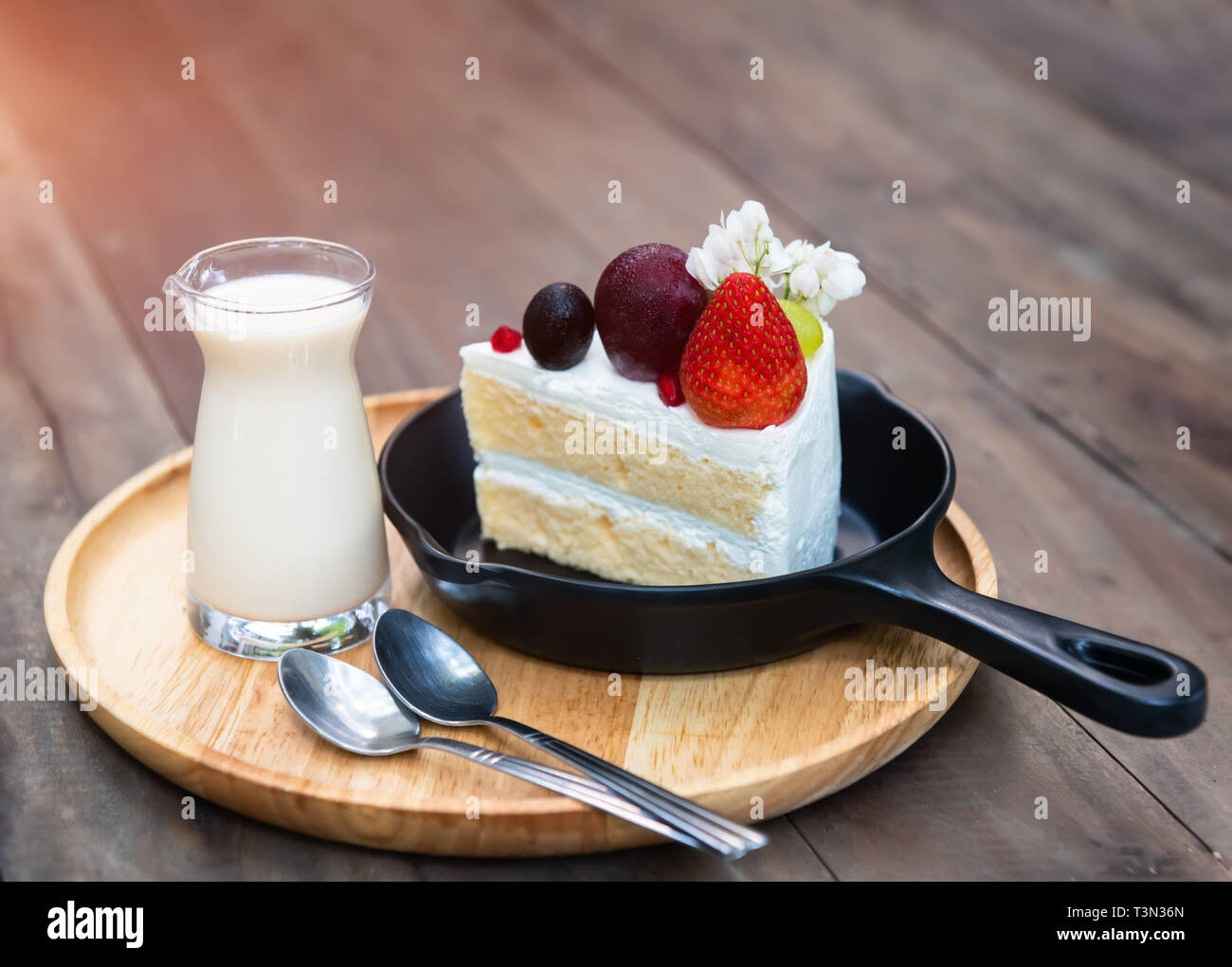 Sweet fruit cream cake cut serve with milk glass set on wooden plate with indoor low lighting. Stock Photo