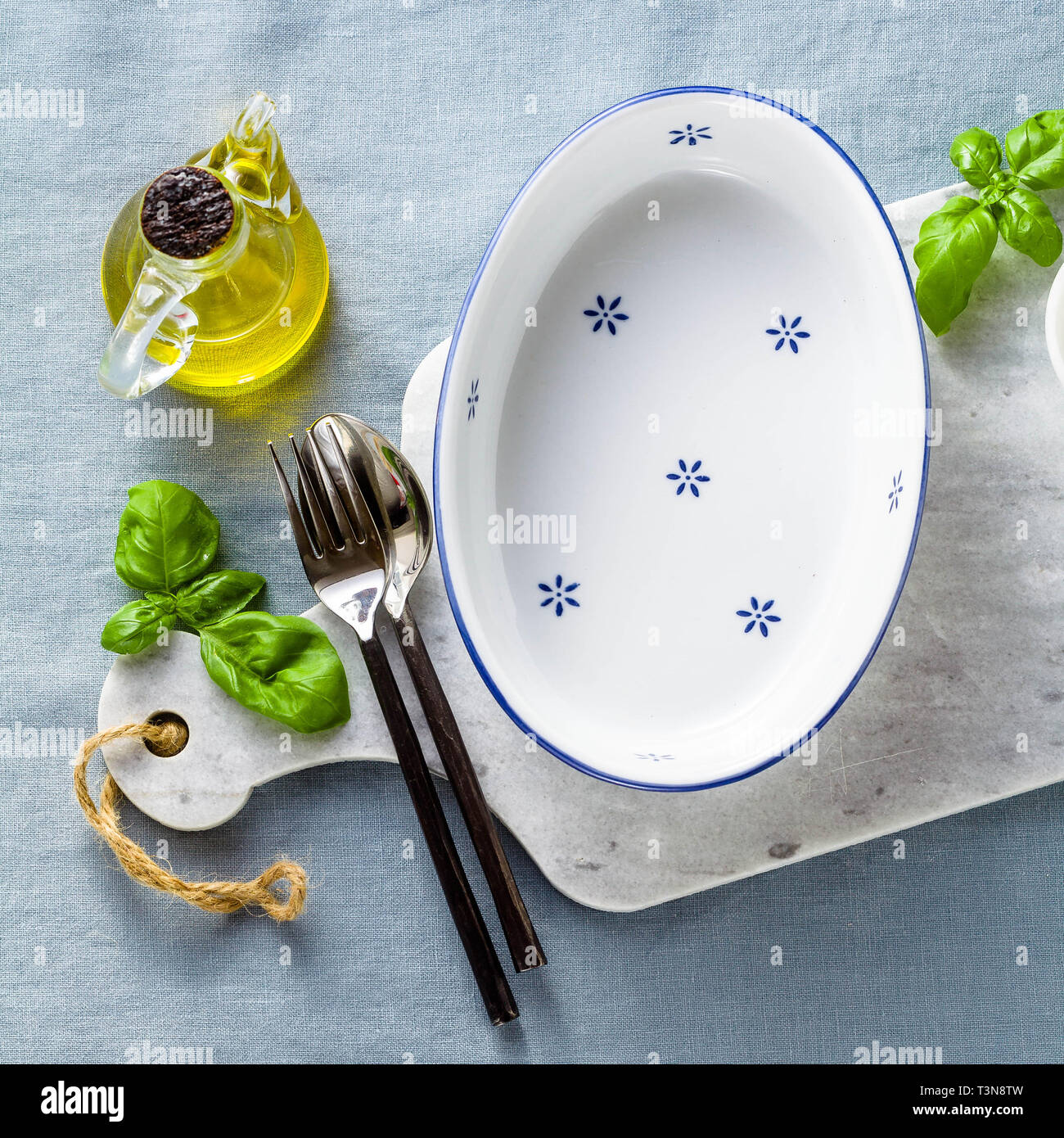 empty ceramic white baking dish on a marble cutting board and cutlery on a blue linen tablecloth with olive oil, spices and basil leaves. home cooking Stock Photo