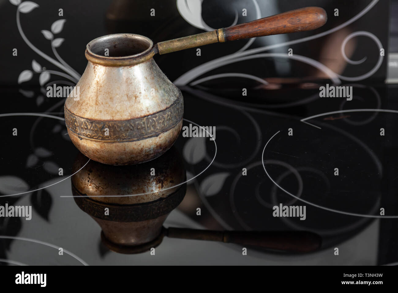 Old Bronze Turkish retro coffee maker kanaka on glass hob and stove with wooden handle, worn on a black background in an apartment in the kitchen. Ret Stock Photo