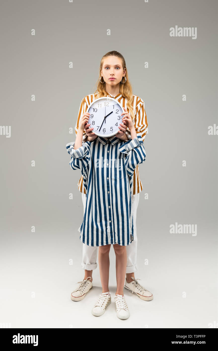 Little girl in oversize striped shirt closing her face with round clock Stock Photo