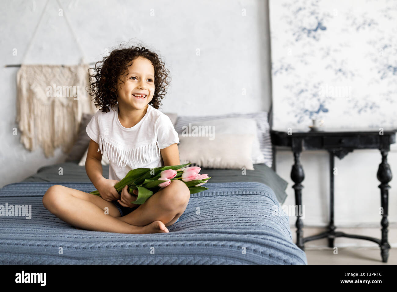 A little pretty curly girl sitting on her bed with tulips in her arms. Stock Photo