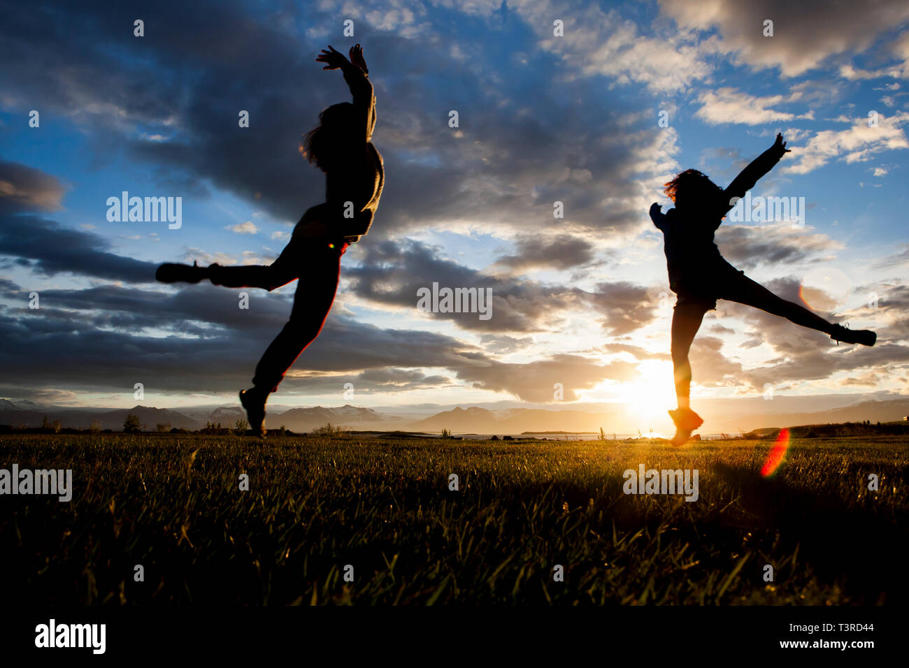 two girls leap through the air backlit by a dramatic mountain sunset in Iceland Stock Photo