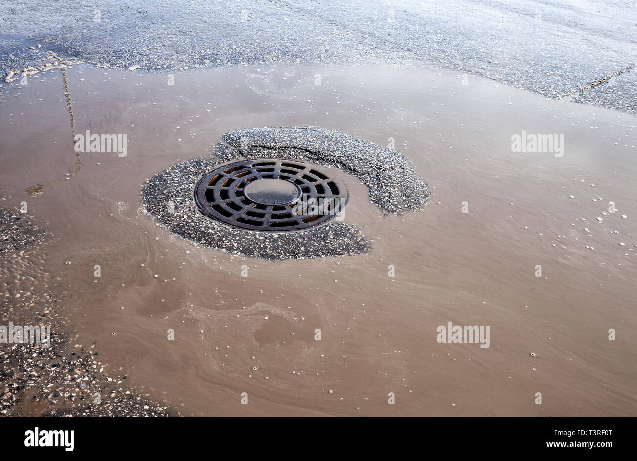 Melted water flows down through the manhole cover on urban asphalt road. Round drainage sewer manhole Stock Photo