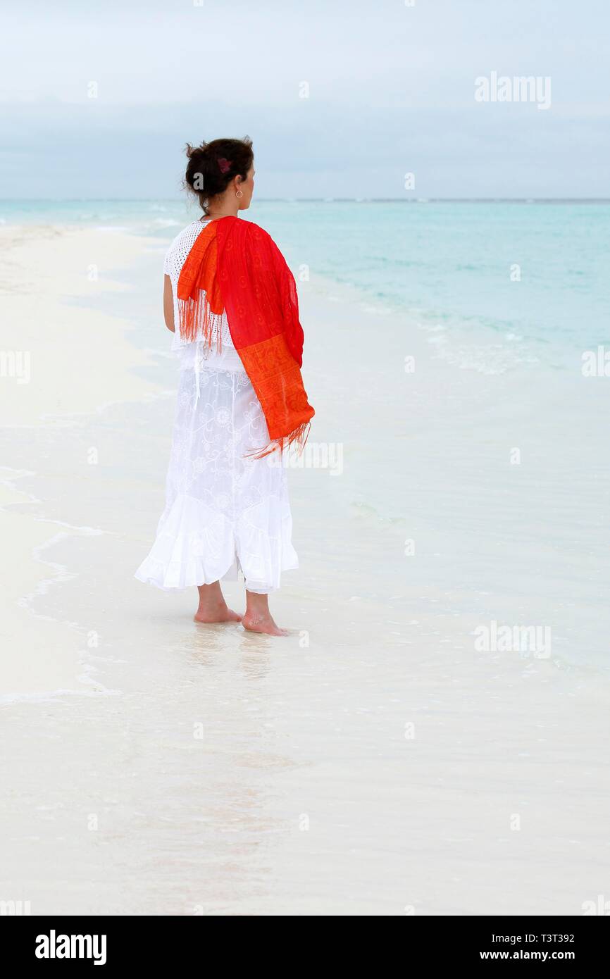Woman with a red scarf standing on a white beach looking at the turquoise blue sea, Maldives Stock Photo
