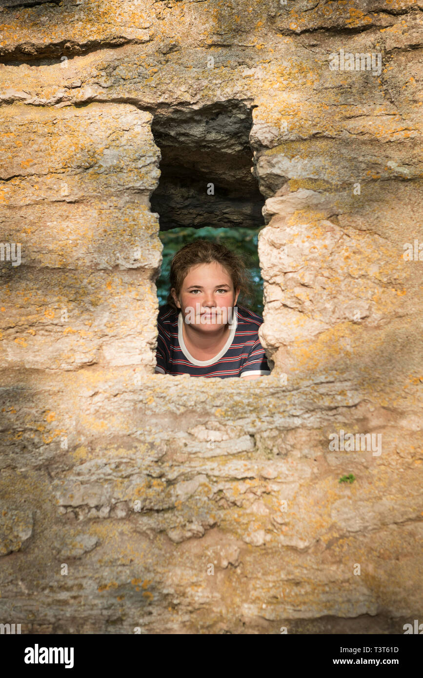 Portrait of a 12 year old girl with brown hair looking at camera through a hole in a stone wall. Stock Photo