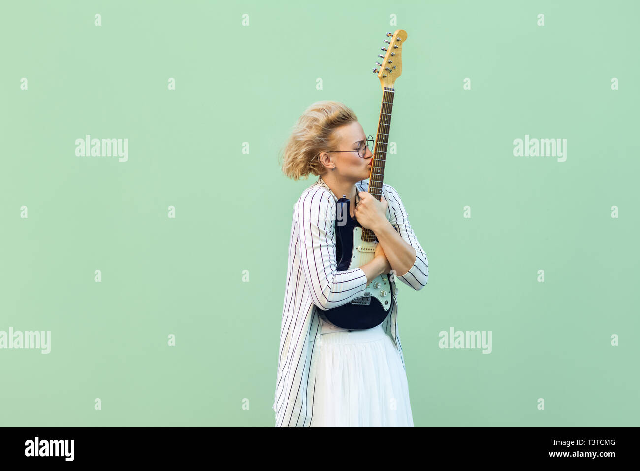 Portrait of young blonde woman in white shirt, skirt, and striped blouse with eyeglasses standing, holding and kissing her lovely electric guitar. ind Stock Photo
