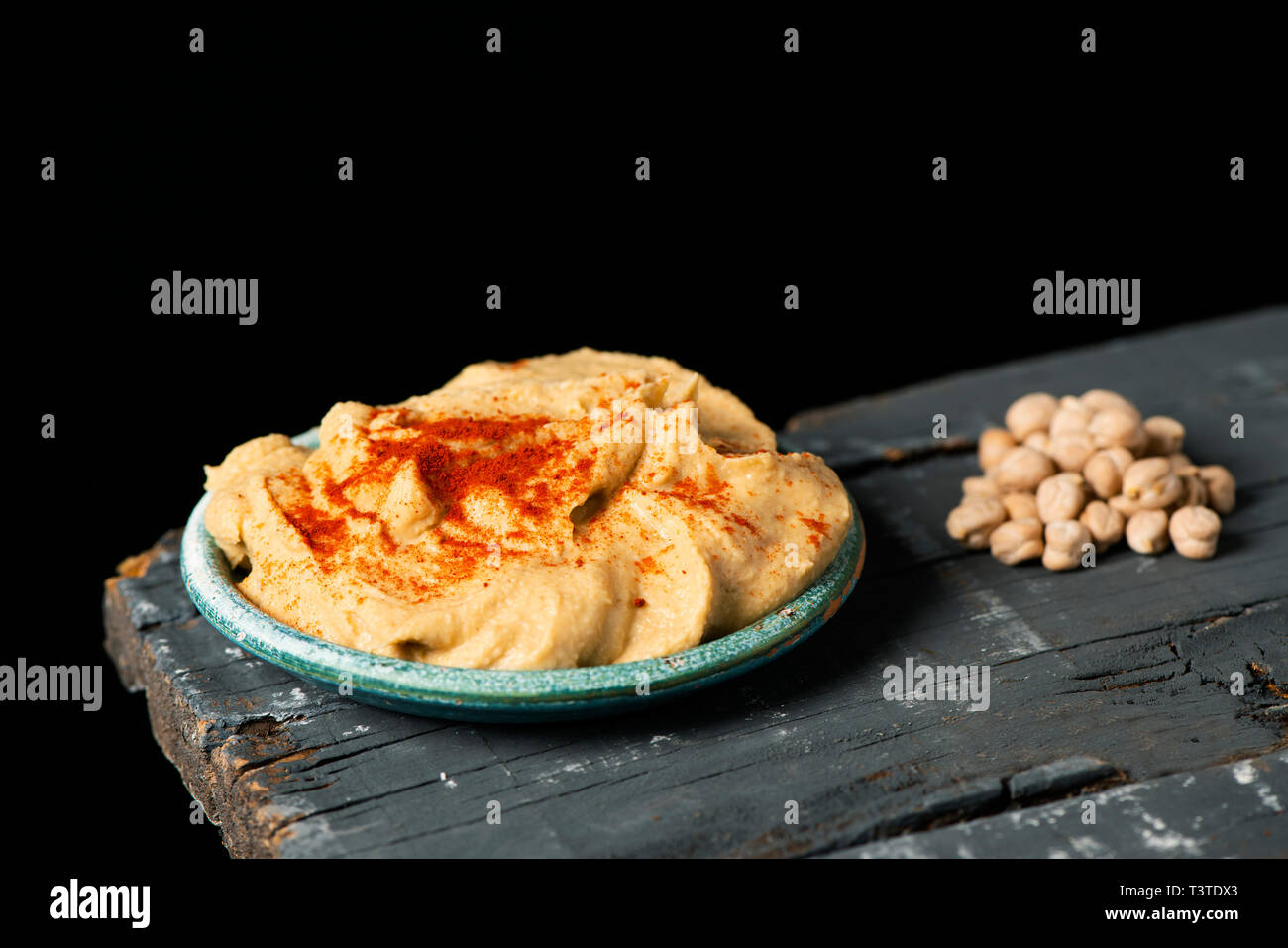 closeup of a green ceramic plate with homemade hummus seasoned with paprika, on a dark gray rustic wooden table against a black background Stock Photo