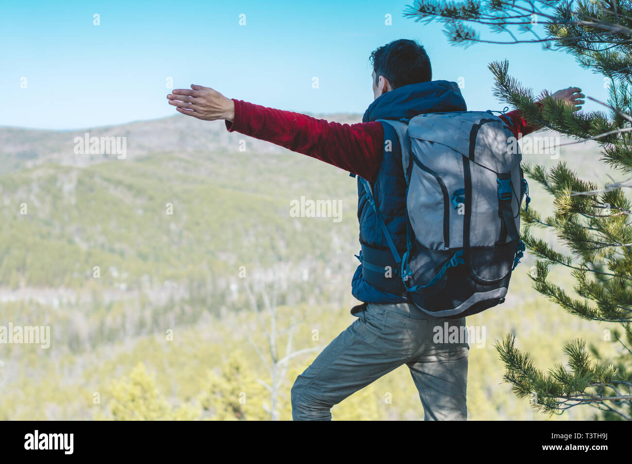 A male tourist hiker with a large backpack stands on a cliff stone in front of a green valley with his arms outstretched. The concept of freedom in tr Stock Photo
