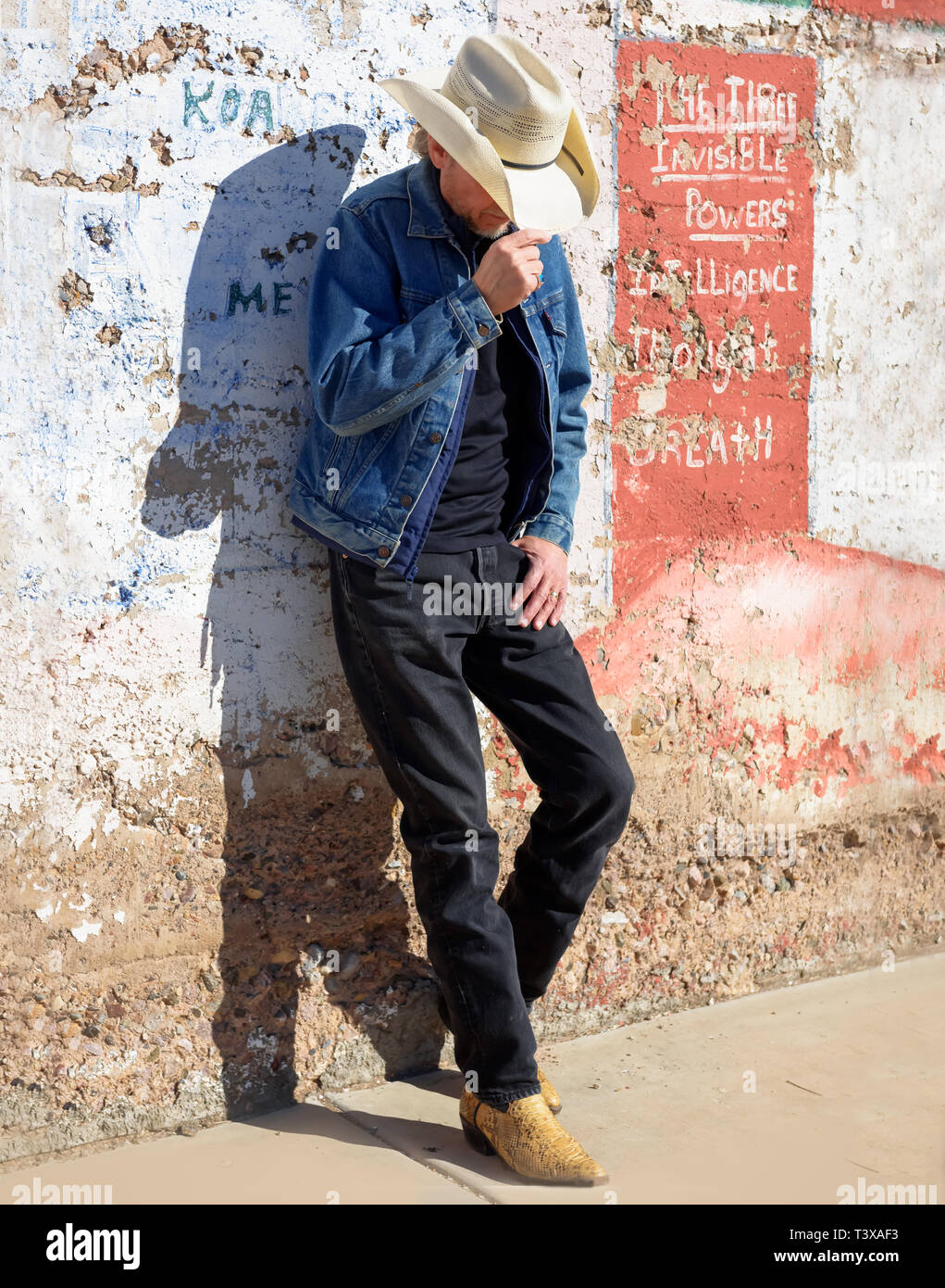 Old cowboy wearing cowboy boots and a hat leans up against a wall in a small southwestern town in Arizona. Stock Photo