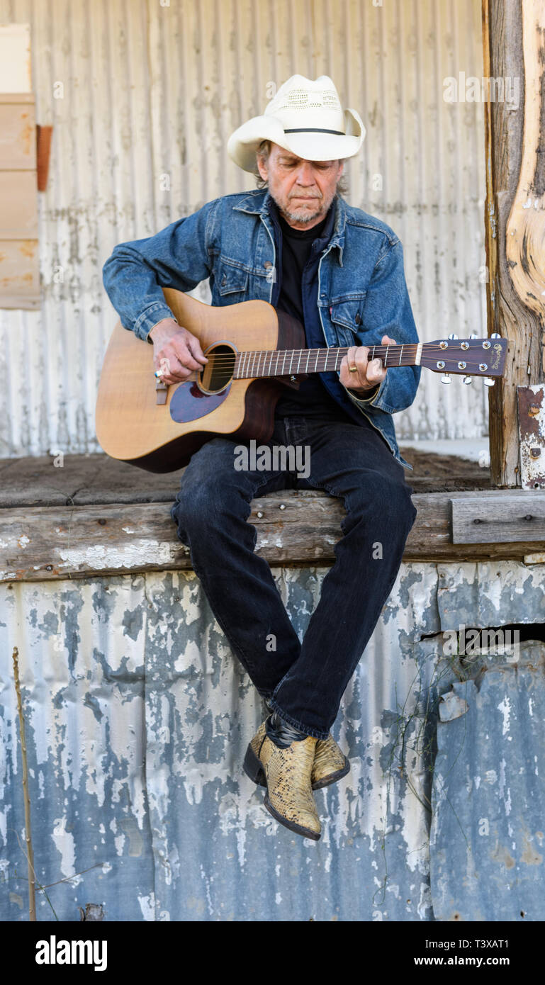 Old Cowboy with cowboy boots strums a guitar while sitting on an abandoned loading dock in rural USA. Stock Photo