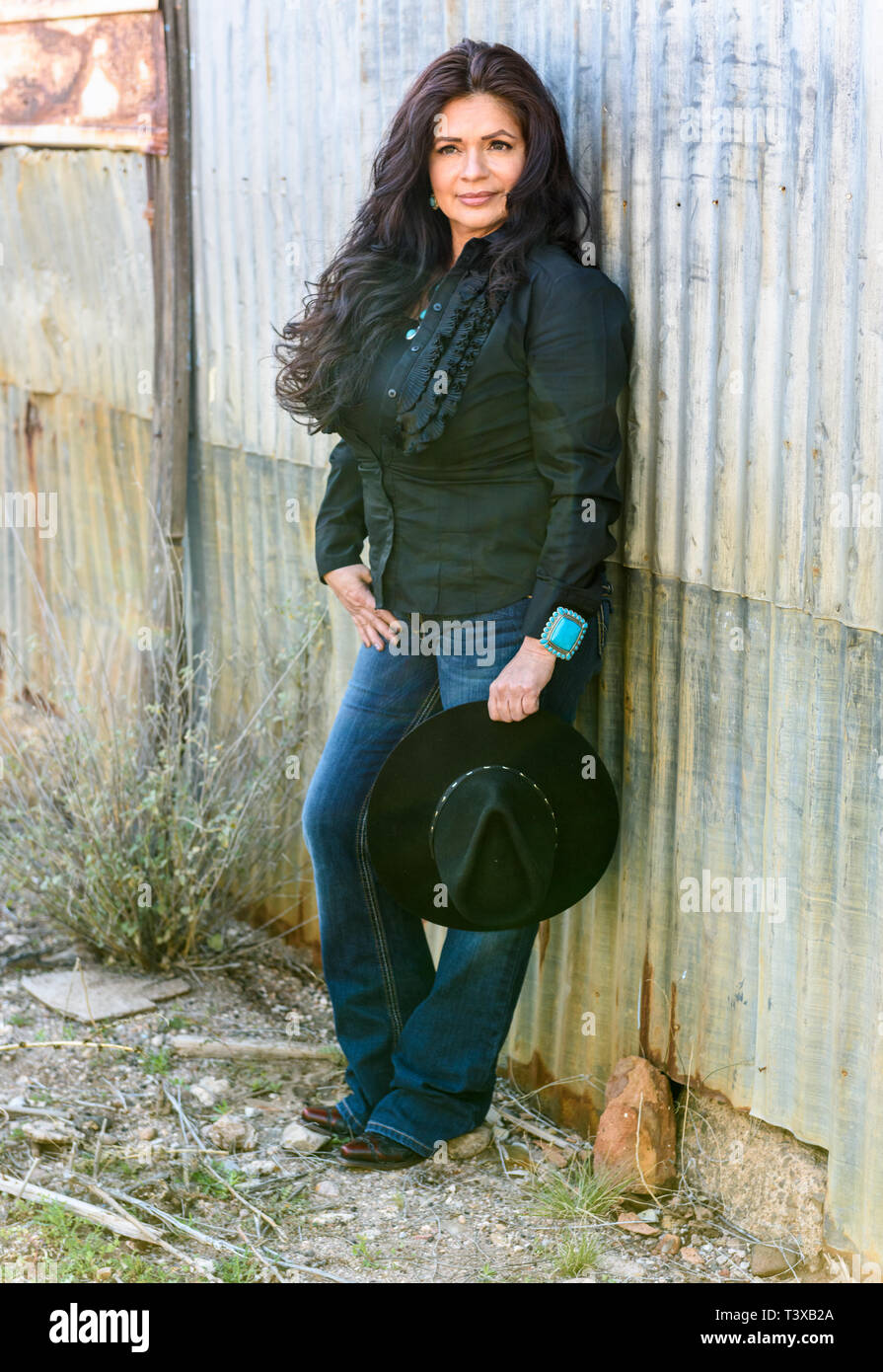Attractive woman with long, dark hair holds cowboy hat in rustic, rural setting. Stock Photo