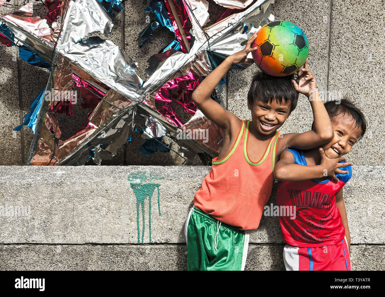 Manila, Philippines: Two young boys holding a football laughing and posing in front of a wall and a broken christmas decoration Stock Photo