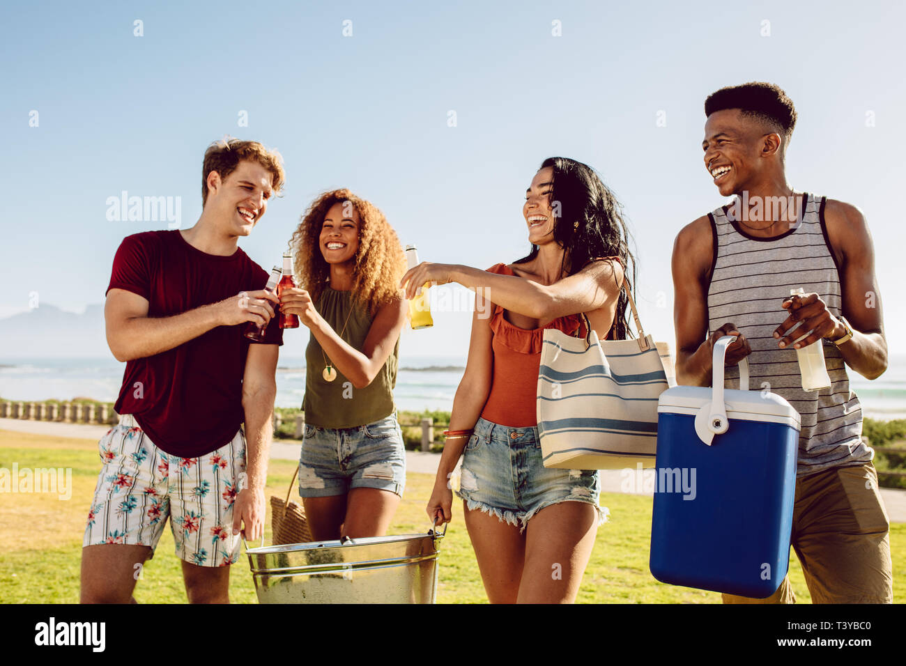 Group of people carrying cooler and beverage tub for party on beach. Diverse group of young people walking outdoors and having drinks. Stock Photo