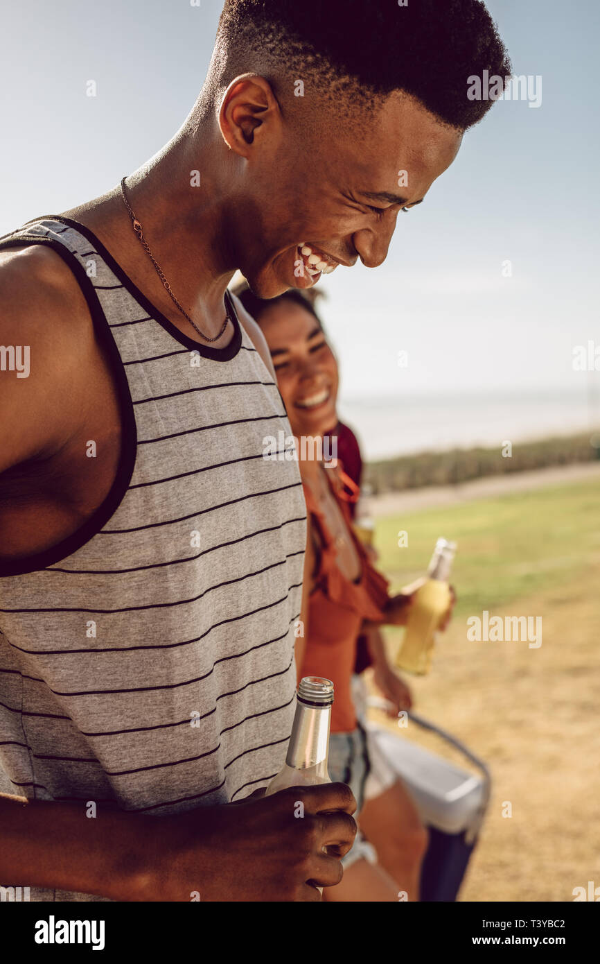 Smiling african man holding beer walking with friends outdoors on summer day. Friends having beers while walking to the picnic spot. Stock Photo