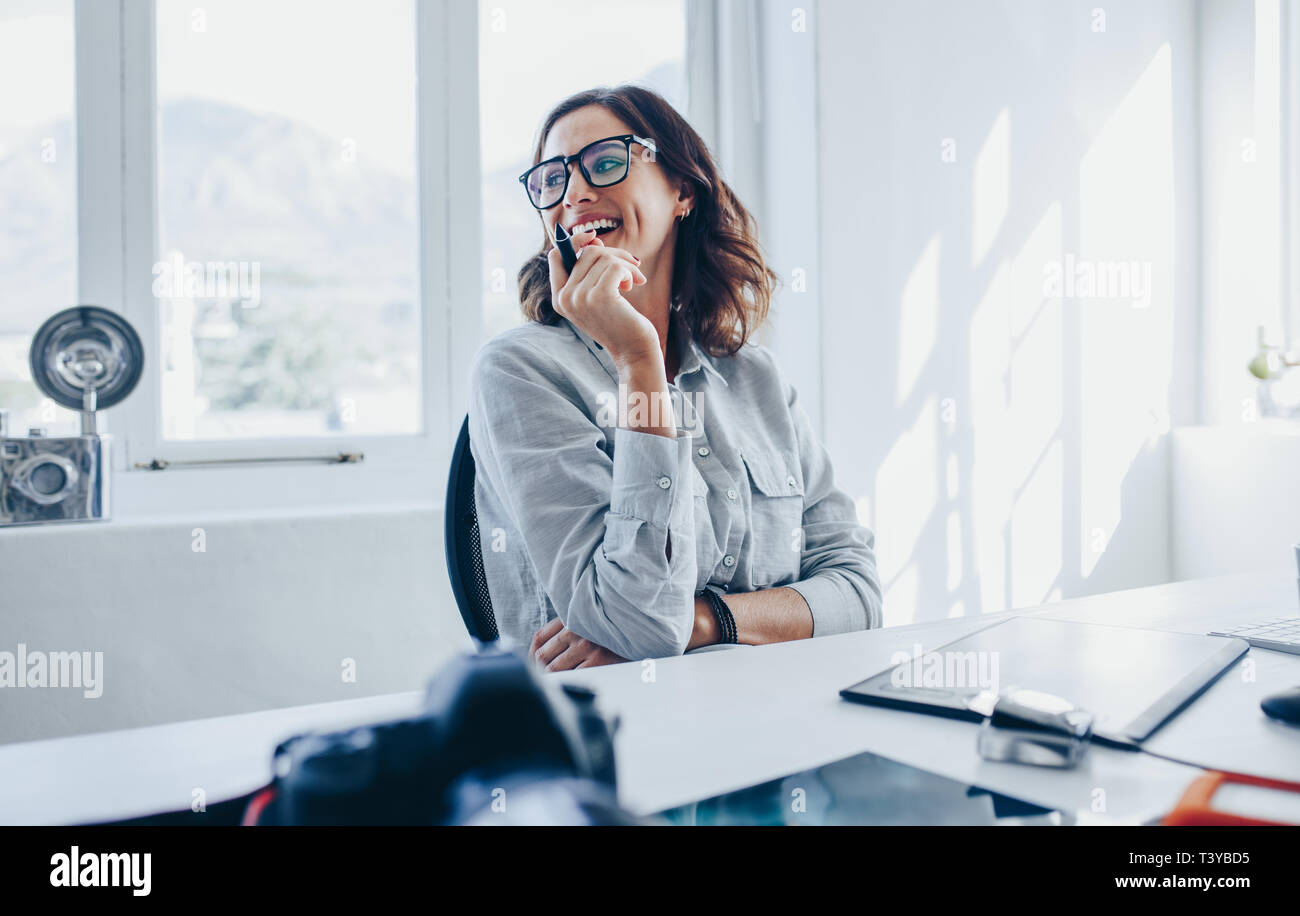 Female creative professional sitting at her desk looking away and smiling. Woman photographer in her office. Stock Photo
