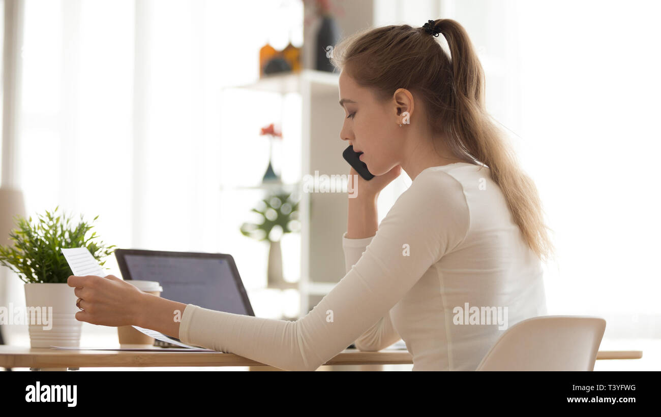 Serious young woman talking on phone, reading paper document Stock Photo
