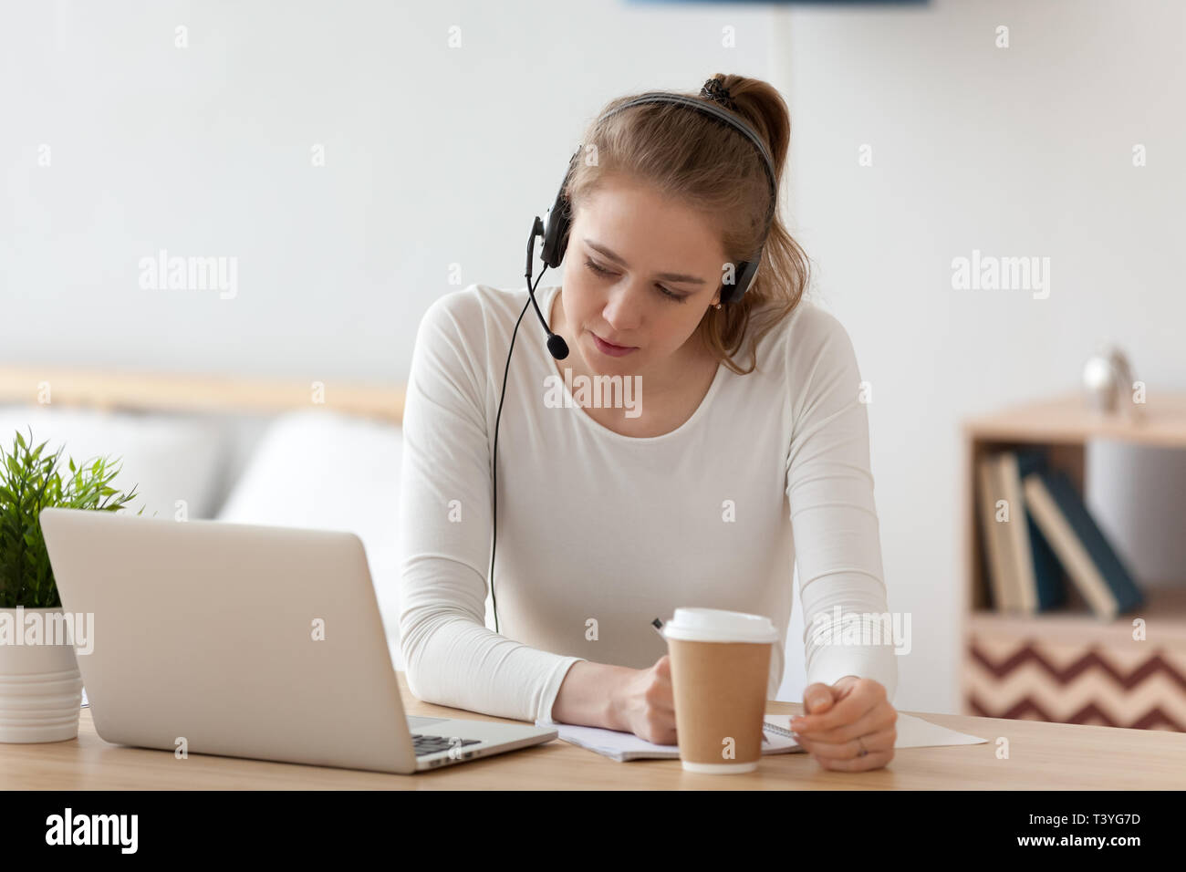 Focused woman in headset using laptop, writing notes Stock Photo