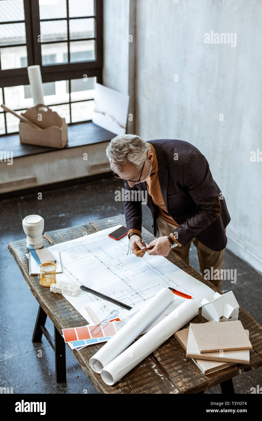 Making some notes. Grey-haired interior designer making some notes on the sketches in his light workshop Stock Photo