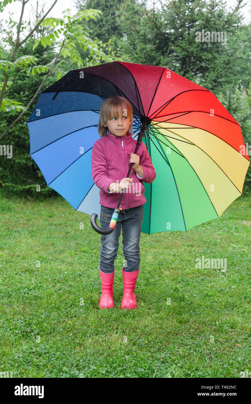 Little girl wearing fleece jacket and rubber boots standing hiding under colorful umbrella in a rain - drizzly weather concept Stock Photo