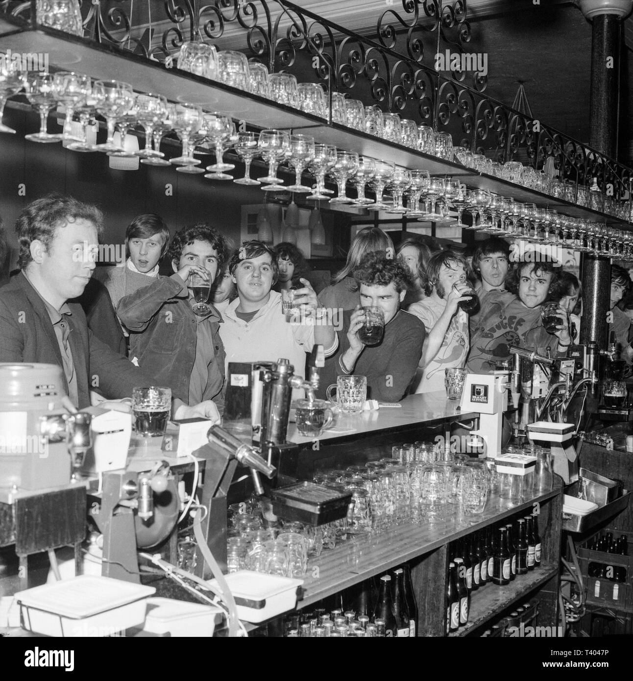 A group of mainly young men standing around  a bar in an English pub during the 1970s, mostly drinking beer. Black and white photograph. Stock Photo