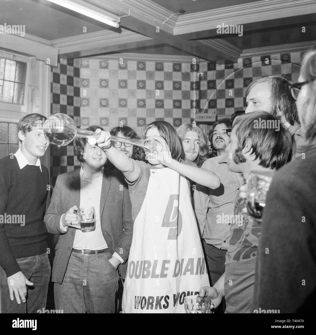 A young man drinking a Yard Of Ale in an English pour during the 1970s.A yard of ale or yard glass is a very tall beer glass used for drinking around 2 1⁄2 imperial pints (1.4 L) of beer, depending upon the diameter. The glass is approximately 1 yard (90 cm) long, shaped with a bulb at the bottom, and a widening shaft, which constitutes most of the height. The glass most likely originated in 17th-century England, where the glass was known also as a 'long glass', a 'Cambridge yard (glass)' and an 'ell glass'. It is associated by legend with stagecoach drivers. Stock Photo