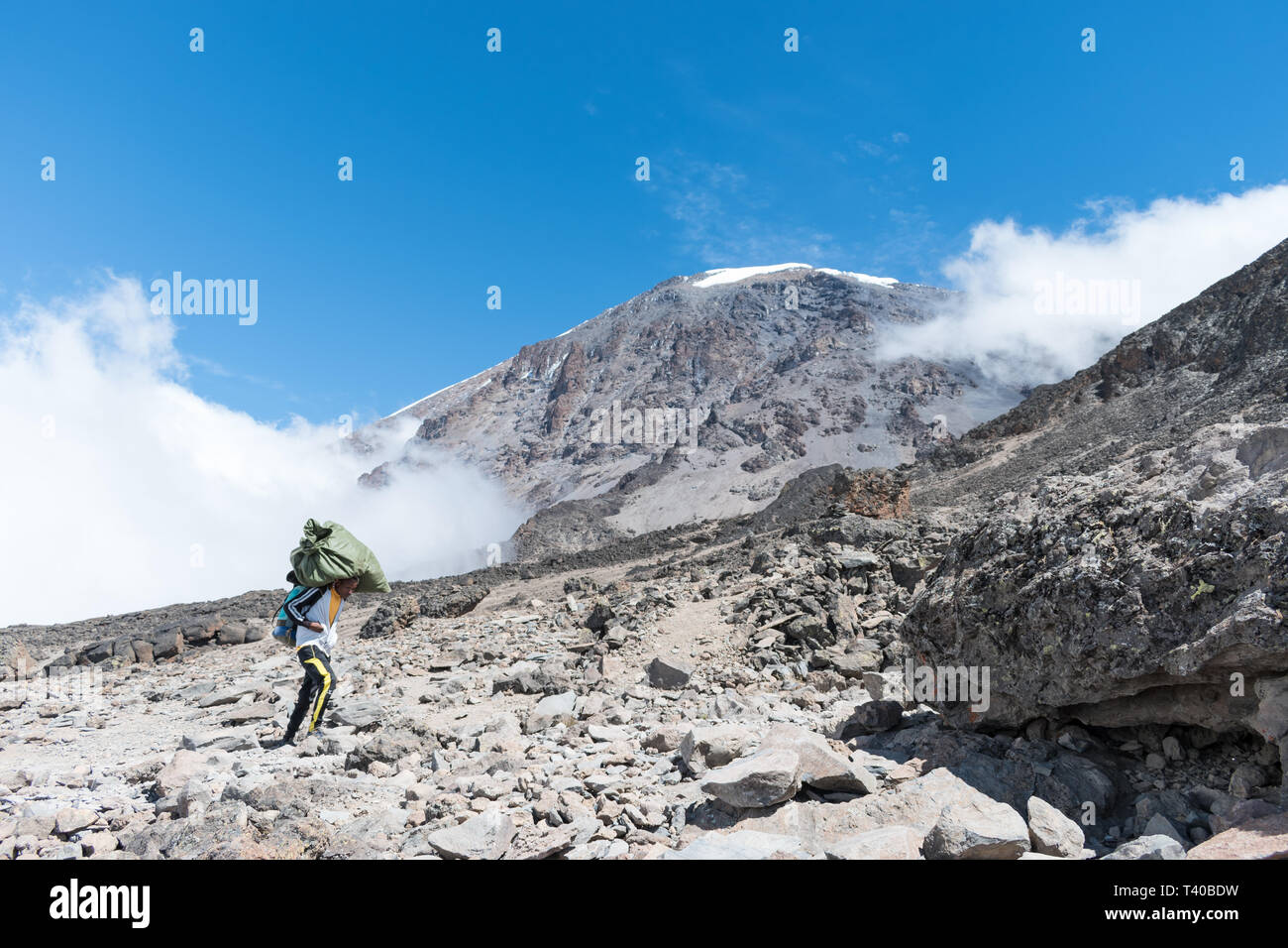 A single male porter carries a heavy load on his back and head in the hot sun on a clear and cloudy day. Summit of Kilimanjaro is in the background. Stock Photo