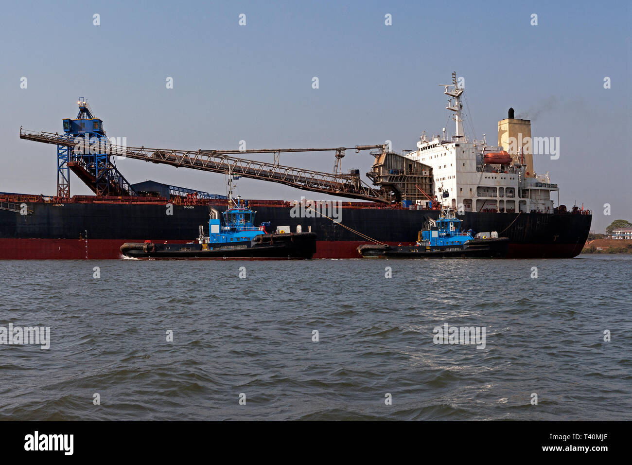 Port operations for managing and transporting iron ore. Transhipping boat and tugs prepare to set sail to unload into OGV - Ocean Going Vessel at sea Stock Photo