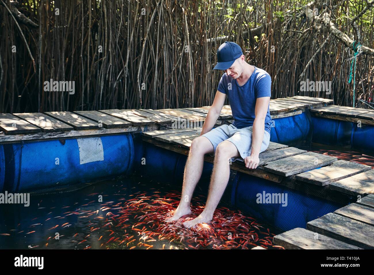 Young man having natural fish spa therapy and foot massage against mangrove trees in lake, Sri Lanka. Stock Photo
