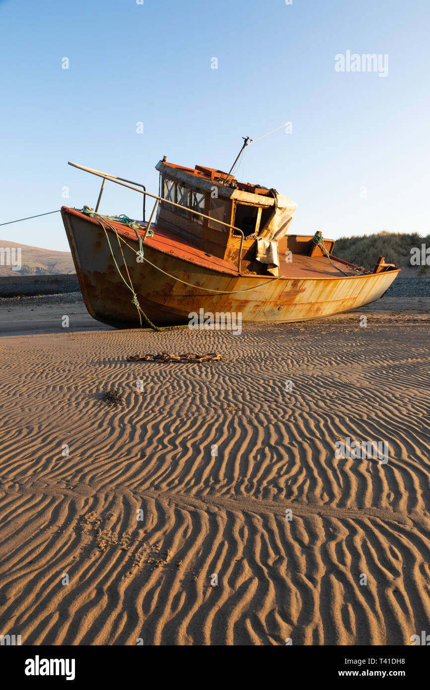 Barmouth, Gwynedd / Wales - April 10th 2019: In the light of the setting sun, a small rusting boat lies on rippled sand after the tide has gone out. Stock Photo