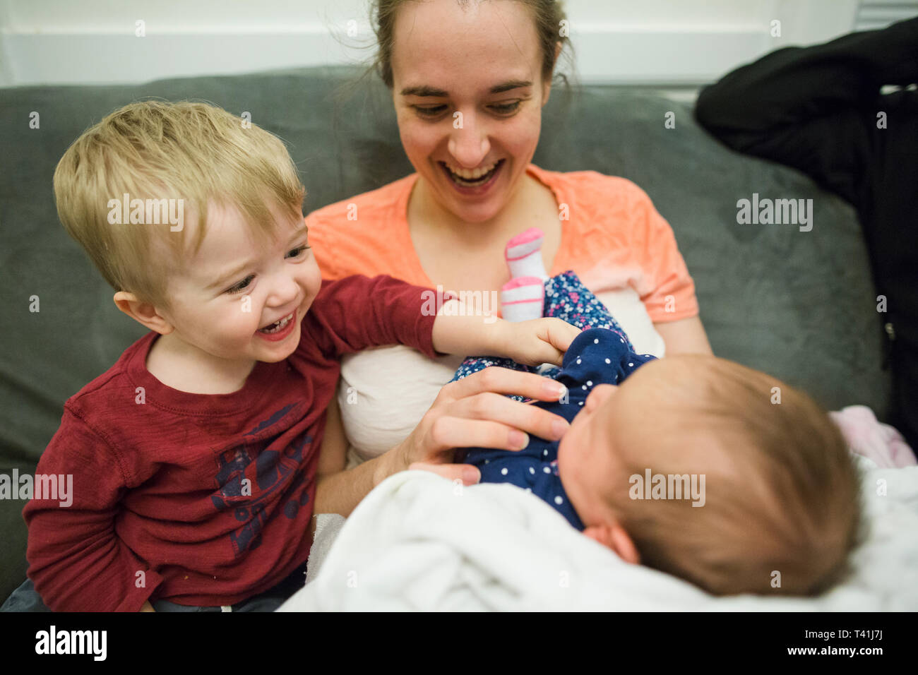 Adorable baby boy smiles at newborn baby girl being held by mother Stock Photo