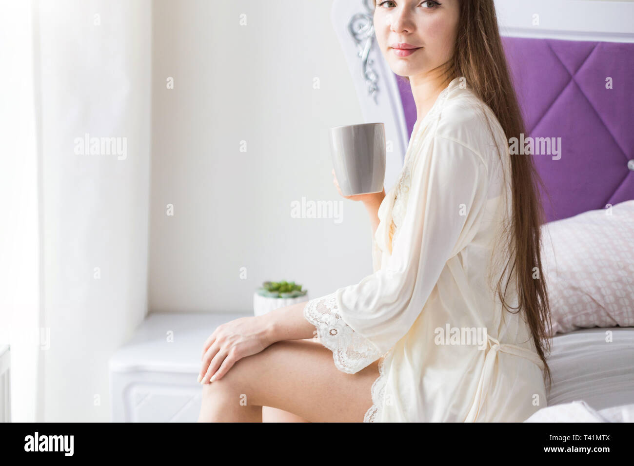 Attractive young girl in nightwear sits on the edge of the bed by the ...