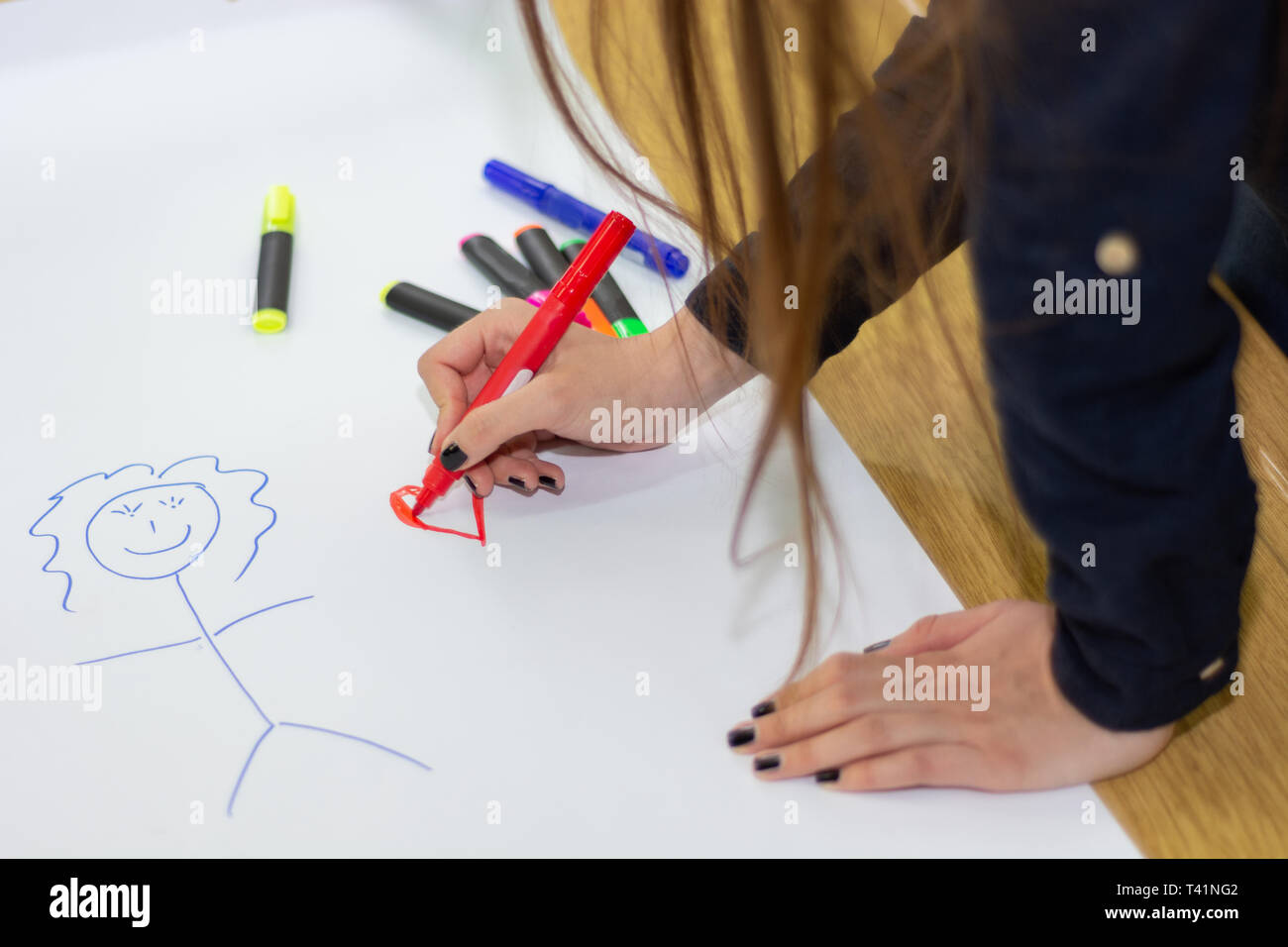 Woman hand with red felt pen drawing heart on big white paper with stickman on desk in classroom. Colorful pens spilled on paper. Education concept Stock Photo