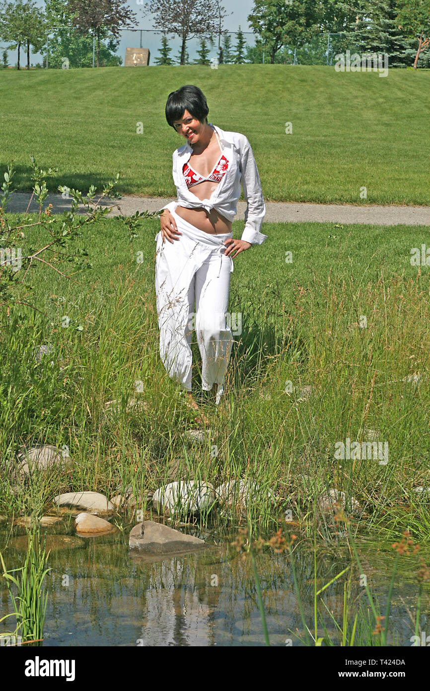 Dark haired beautiful woman posing In a park wearing a red and white bikini top, white jacket and pants Stock Photo