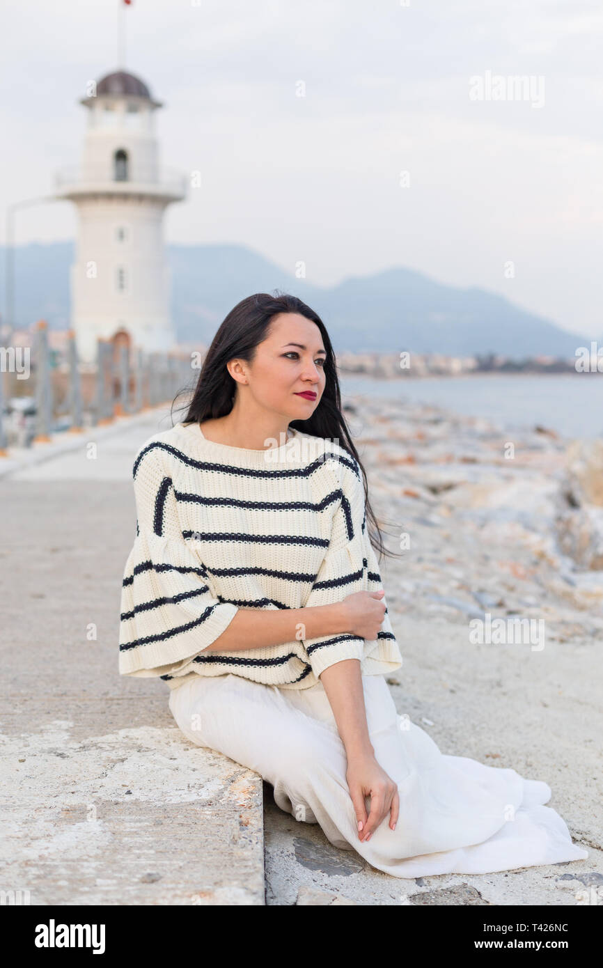 Brunette female dressed in nautical fashion sitting on the rocks with  lighthouse and mountains blurred as background. Vertical composition Stock  Photo - Alamy