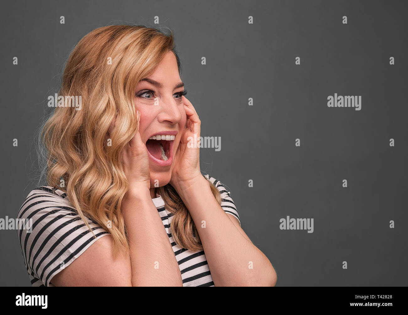 Emotional blonde screams. Frightened woman screaming straining standing on a gray background. Stock Photo