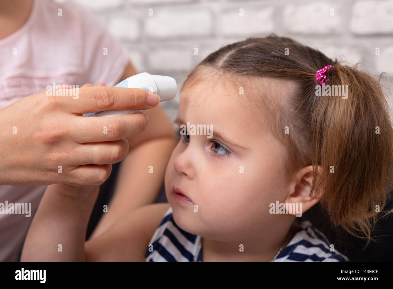 Mother Checking Fever Of Her Sick Daughter Using Forehead Thermometer At Home Stock Photo