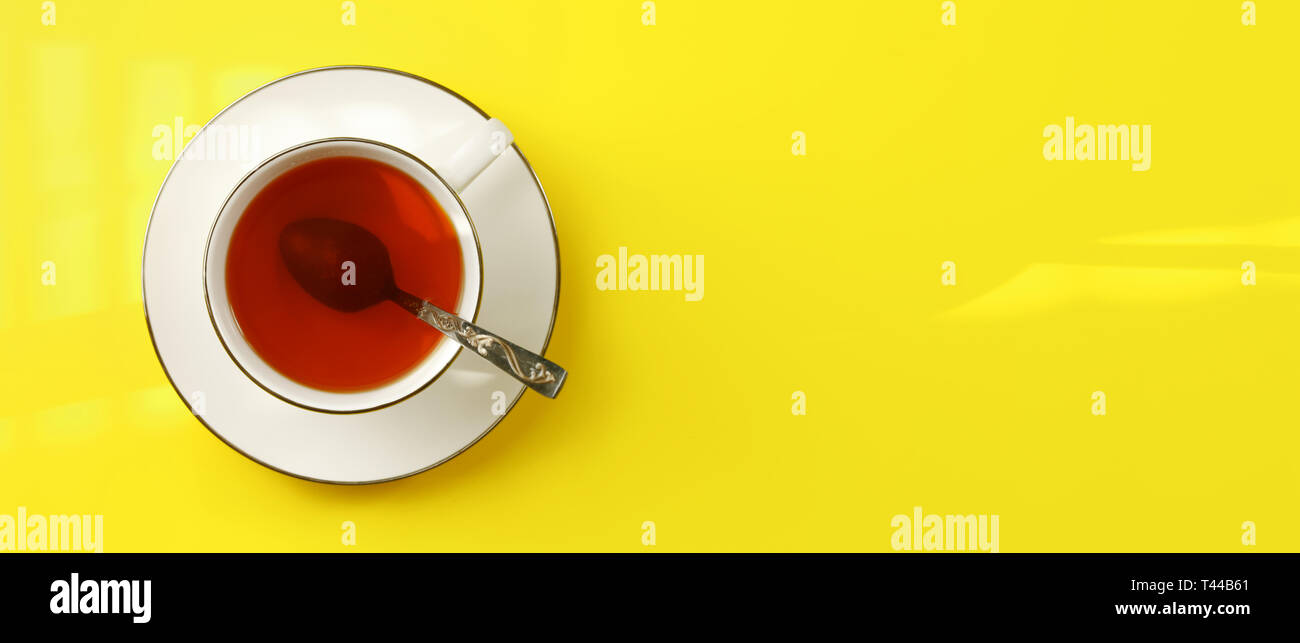 Flat lay photo - white porcelain cup with spoon filled hot amber tea, on yellow board, wide banner space for text right Stock Photo
