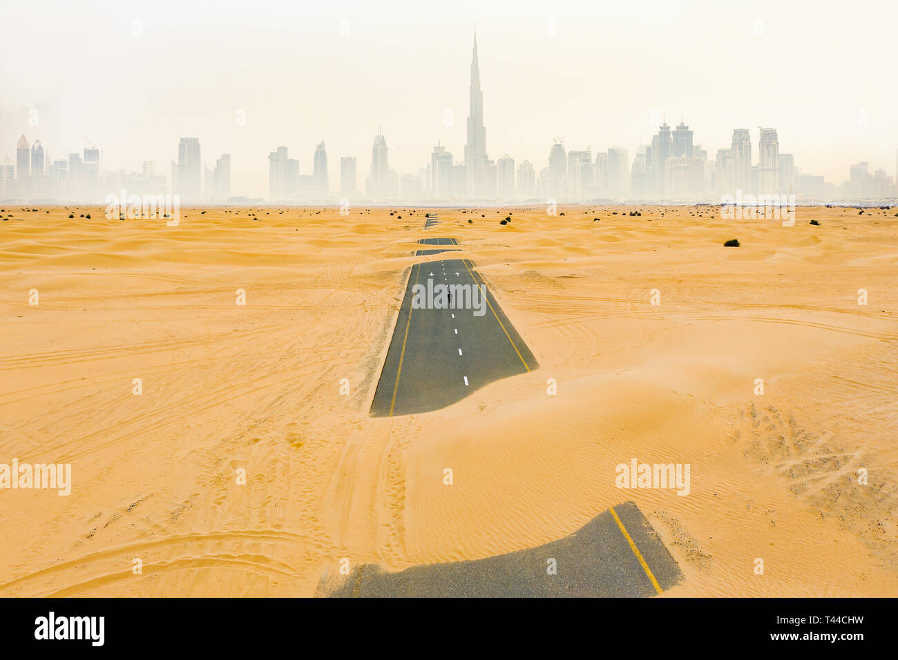 Stunning aerial view of an unidentified person walking on a deserted road covered by sand dunes in Dubai desert. Dubai skyline surrounded by fog Stock Photo
