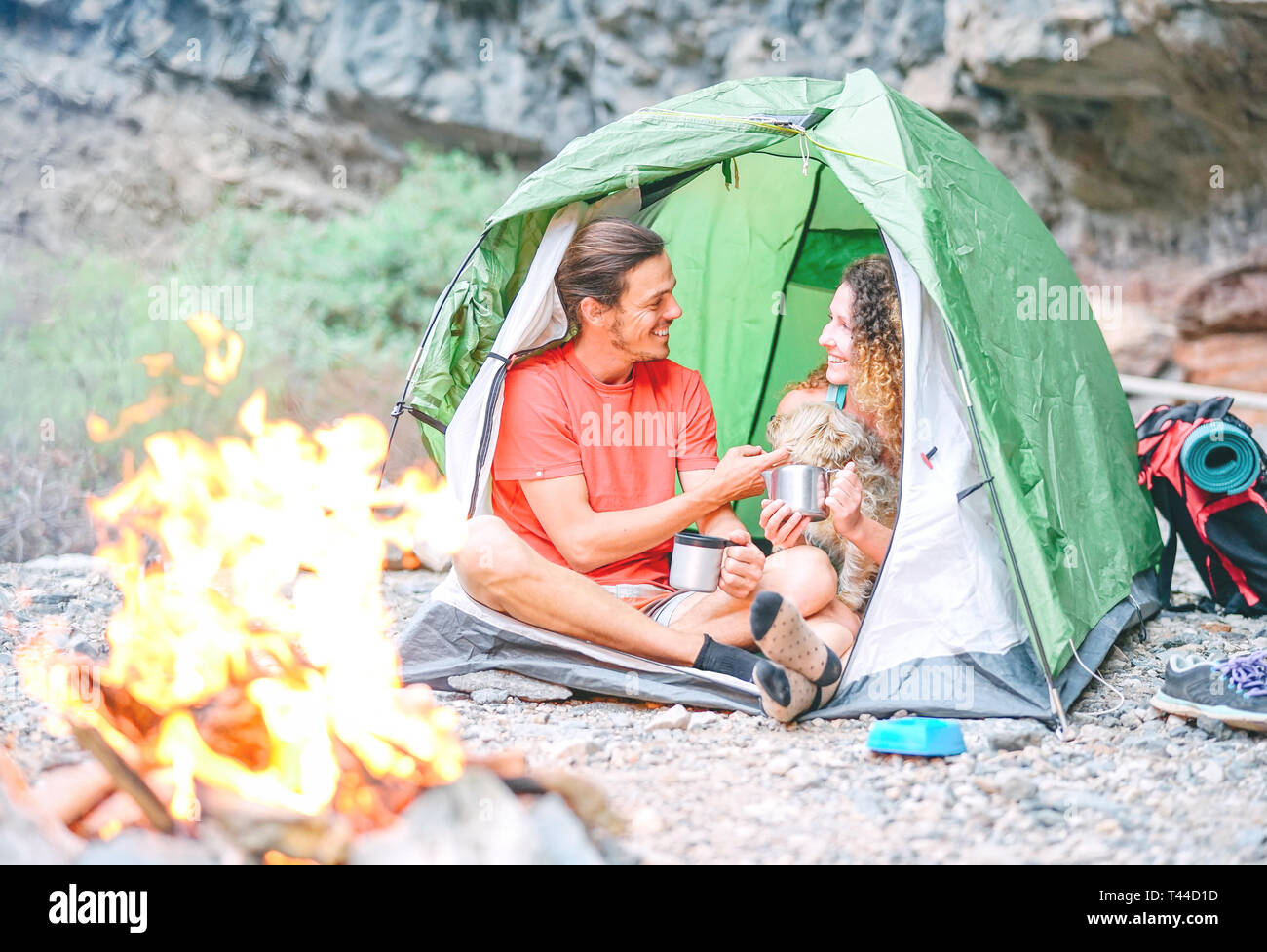 Happy couple of hikers with their dog camping with tent around rock mountains next to fire - People relaxing in a camp drinking hot tea Stock Photo