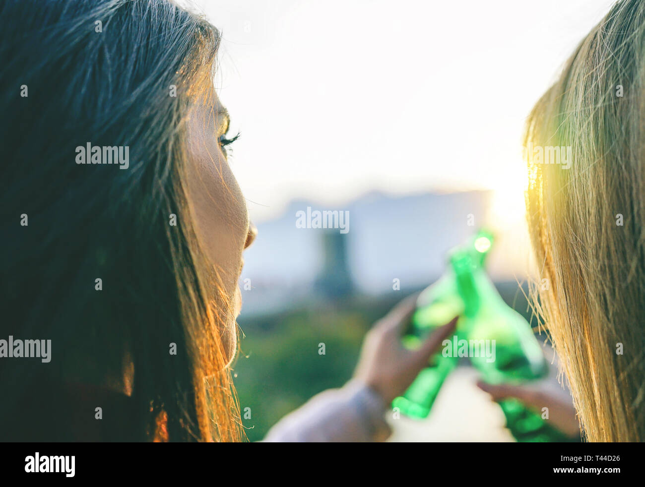 Young women toasting beer on a sunset - Happy friends drinking and cheering beers in a park outdoor admiring the beautiful view Stock Photo