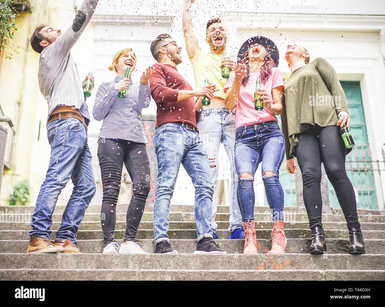 Group of happy friends celebrating together throwing up confetti and drinking beers - Young people having a party on stairs of an urban area Stock Photo
